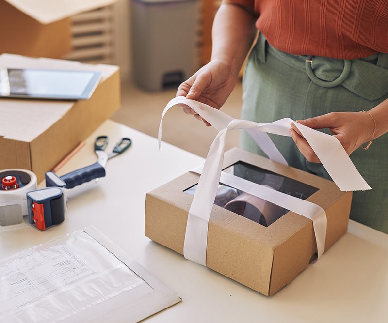 close up woman decorating cardboard box with ribbon table before delivery